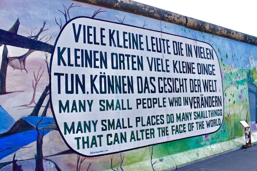 A graffiti mural on a large wall with a message written in German and English. The German text reads, 'Viele kleine Leute die in vielen kleinen Orten viele kleine Dinge tun, können das Gesicht der Welt,' which translates to the English text below it, 'Many small people who in many small places do many small things that can alter the face of the world.' The mural features a colorful background with abstract images of trees and landscapes.