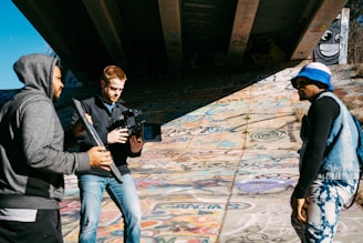 Three individuals are engaged in what appears to be a film or photography session under a bridge. The person on the left is holding a reflector, and the person in the center is operating a large camera rig. The individual on the right is wearing a blue hat and denim vest, standing on a graffiti-covered surface. Sunlight casts shadows across the scene.