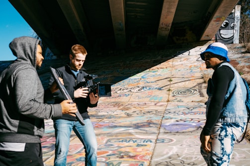 Three individuals are engaged in what appears to be a film or photography session under a bridge. The person on the left is holding a reflector, and the person in the center is operating a large camera rig. The individual on the right is wearing a blue hat and denim vest, standing on a graffiti-covered surface. Sunlight casts shadows across the scene.