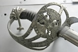 An intricately designed metal sword hilt with ornate carvings and a depiction of an eagle holding a shield. The handle is wrapped in a dark material, and the metal has a polished, silver appearance.