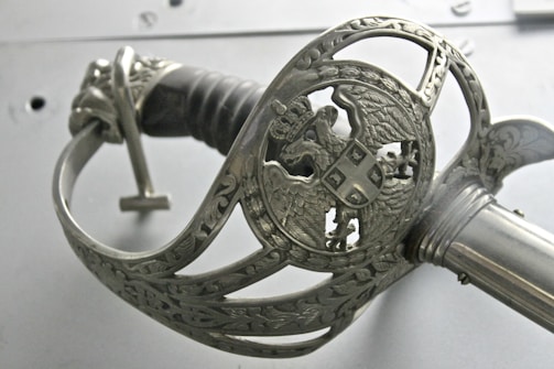An intricately designed metal sword hilt with ornate carvings and a depiction of an eagle holding a shield. The handle is wrapped in a dark material, and the metal has a polished, silver appearance.
