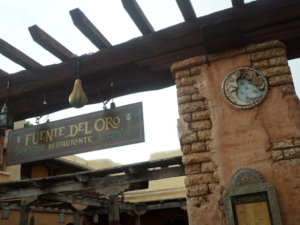 A rustic restaurant sign labeled 'Fuente Del Oro Restaurante' hangs from a wooden beam. The structure features weathered wooden elements and a stone wall with a decorative sun or moon emblem. The color palette is earthy, with shades of brown and beige.