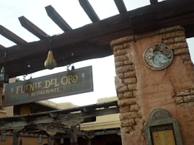 A rustic restaurant sign labeled 'Fuente Del Oro Restaurante' hangs from a wooden beam. The structure features weathered wooden elements and a stone wall with a decorative sun or moon emblem. The color palette is earthy, with shades of brown and beige.