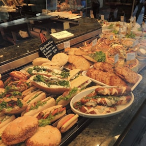 A display of various freshly prepared sandwiches and baked goods, including baguettes filled with lettuce and vegetables, bread rolls, and pretzel sandwiches. The food items are neatly arranged in a glass counter and labeled with small signs indicating their names and prices. There is a selection of flatbreads and pastries placed alongside garnished with fresh herbs.