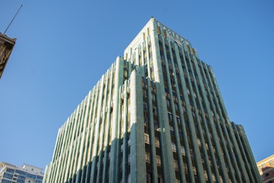 A tall, green Art Deco skyscraper reaches towards a clear blue sky, with sunlight casting shadows along its intricate architectural details. Its stylized design features vertical lines and decorative elements typical of the early 20th century.