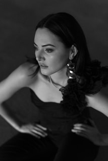 A monochrome portrait of a woman gazing thoughtfully to the side, wearing a single-shoulder dress with ruffled details and large circular earrings. Her hands are placed on her hips.