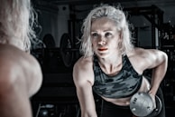 A woman with blonde hair is engaged in a workout, lifting a dumbbell with a focused expression. She is wearing a camouflage sports bra, and her muscles are visibly toned. The setting is a dimly lit gym with various workout equipment visible in the background.