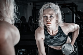 A woman with blonde hair is engaged in a workout, lifting a dumbbell with a focused expression. She is wearing a camouflage sports bra, and her muscles are visibly toned. The setting is a dimly lit gym with various workout equipment visible in the background.