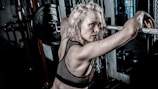 A woman with light blonde hair is in a gym setting, leaning against a barbell on a weightlifting rack. She has an intense and focused expression, wearing a black mesh sports bra. The gym environment includes weight plates and chains, suggesting a strength training area.