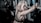 A woman with light blonde hair is in a gym setting, leaning against a barbell on a weightlifting rack. She has an intense and focused expression, wearing a black mesh sports bra. The gym environment includes weight plates and chains, suggesting a strength training area.