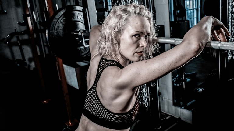 A woman with light blonde hair is in a gym setting, leaning against a barbell on a weightlifting rack. She has an intense and focused expression, wearing a black mesh sports bra. The gym environment includes weight plates and chains, suggesting a strength training area.