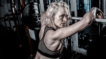 A woman with light blonde hair is in a gym setting, leaning against a barbell on a weightlifting rack. She has an intense and focused expression, wearing a black mesh sports bra. The gym environment includes weight plates and chains, suggesting a strength training area.