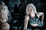A person with blonde hair is exercising in a gym, lifting dumbbells while wearing a camouflage-patterned tank top. The gym setting includes weights and equipment in the background, with dim lighting creating a focused atmosphere.