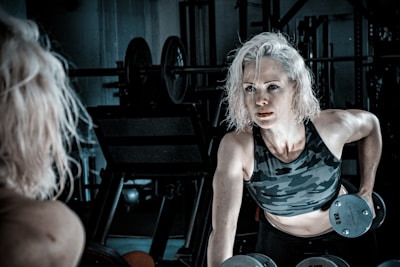 A person with blonde hair is exercising in a gym, lifting dumbbells while wearing a camouflage-patterned tank top. The gym setting includes weights and equipment in the background, with dim lighting creating a focused atmosphere.