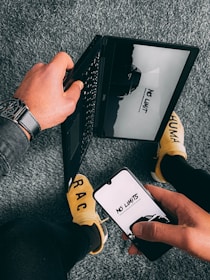 A person is holding a smartphone displaying the words 'NO LIMITS' against a monochrome background. The same message is mirrored on a laptop screen. The person is wearing a watch and sitting on a gray carpeted floor, dressed in dark pants and yellow socks with the letters 'RAC' visible.