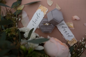 An elegant arrangement featuring a velvet hexagonal ring box containing a delicate gold ring with a small gemstone, surrounded by soft petals and greenery. The background includes calligraphy name tags with gold detailing and scattered rose petals, creating a romantic and sophisticated atmosphere.
