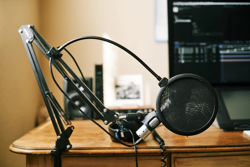 Boom-mounted microphone behind a circular pop filter, with headphones on a wooden desk beside a computer