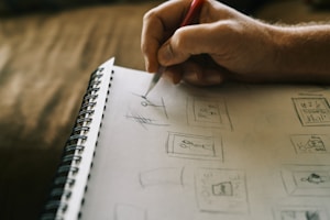 A person's hand is sketching designs on a spiral-bound notebook. The sketches appear to be simple line drawings, including figures and geometric shapes. The focus is on the hand holding a pencil, suggesting an artistic or design process in progress.
