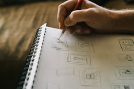 A person's hand is sketching designs on a spiral-bound notebook. The sketches appear to be simple line drawings, including figures and geometric shapes. The focus is on the hand holding a pencil, suggesting an artistic or design process in progress.