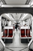 The interior of a modern tram or subway car features rows of red upholstered seats with a gray floor and overhead lighting. There are a few passengers sitting and standing, creating a calm and quiet atmosphere. The design is sleek and minimalist, with stainless steel handles and white paneling.