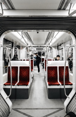 The interior of a modern tram or subway car features rows of red upholstered seats with a gray floor and overhead lighting. There are a few passengers sitting and standing, creating a calm and quiet atmosphere. The design is sleek and minimalist, with stainless steel handles and white paneling.