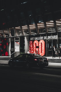 The entrance of the Art Gallery of Ontario is prominent, featuring large illuminated letters 'AGO' with a polka dot pattern. A car is parked on the street in front, and a few people are visible near the entrance. The scene is captured at night, showcasing a modern, urban atmosphere.