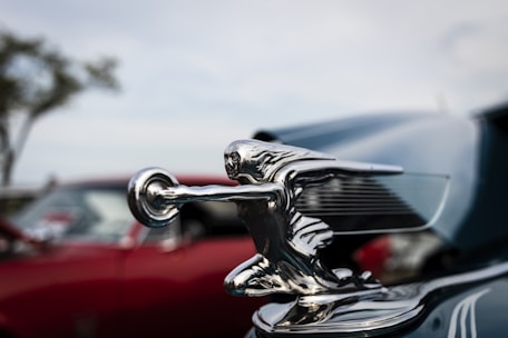 A shiny silver vintage car hood ornament depicting a stylized human figure in a dynamic pose, set against a background of blurred classic cars and a softly out-of-focus tree.