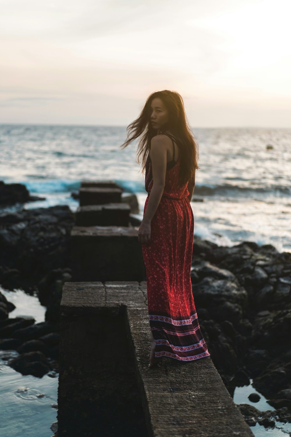 woman in red spaghetti strap dress standing on rock near sea during daytime