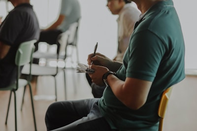 A group of people seated in folding chairs, with some taking notes. The focus is on a person in the foreground, wearing a dark green polo shirt, writing on a notepad with a pen. Other individuals in the background appear slightly blurred, and the environment resembles a meeting or seminar room.