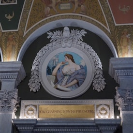 An ornate architectural design featuring a classical figure in a round frame adorned with detailed carvings and floral motifs. A medallion portrays a woman in classical attire, with the words 'How Charming is Divine Philosophy' inscribed in gold lettering beneath.