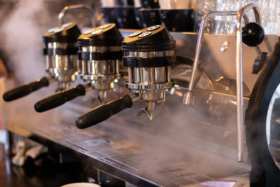 The image shows a stainless steel espresso machine with three black handles and silver knobs, all emitting steam from their spouts. The machine is situated on a kitchen countertop, which also has two additional coffee cups placed nearby.