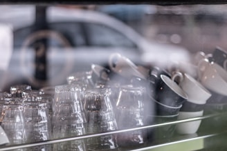 A collection of glass cups and ceramic mugs are neatly arranged on a shelf behind a reflective glass window, with a blurred background that includes a hint of a car. The lighting is soft, giving a calm ambiance.