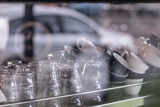 A collection of glass cups and ceramic mugs are neatly arranged on a shelf behind a reflective glass window, with a blurred background that includes a hint of a car. The lighting is soft, giving a calm ambiance.