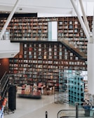A modern library interior featuring a large, well-organized bookshelf wall filled with books. The space has a bright and open design with white walls and floors. A staircase with clear glass railings runs alongside the shelves, and there are contemporary seating arrangements with wooden chairs. Metal frameworks, possibly for display or structural support, are present throughout the area.