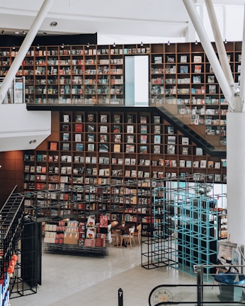 A modern library interior featuring a large, well-organized bookshelf wall filled with books. The space has a bright and open design with white walls and floors. A staircase with clear glass railings runs alongside the shelves, and there are contemporary seating arrangements with wooden chairs. Metal frameworks, possibly for display or structural support, are present throughout the area.