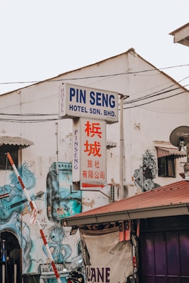 An urban street scene featuring an old, weathered building with a prominent hotel sign in both English and Chinese. The facade is adorned with artistic murals depicting abstract figures. Various signs and electrical wires crisscross the area, and a roof with a Malaysian flag draped over the side is visible.