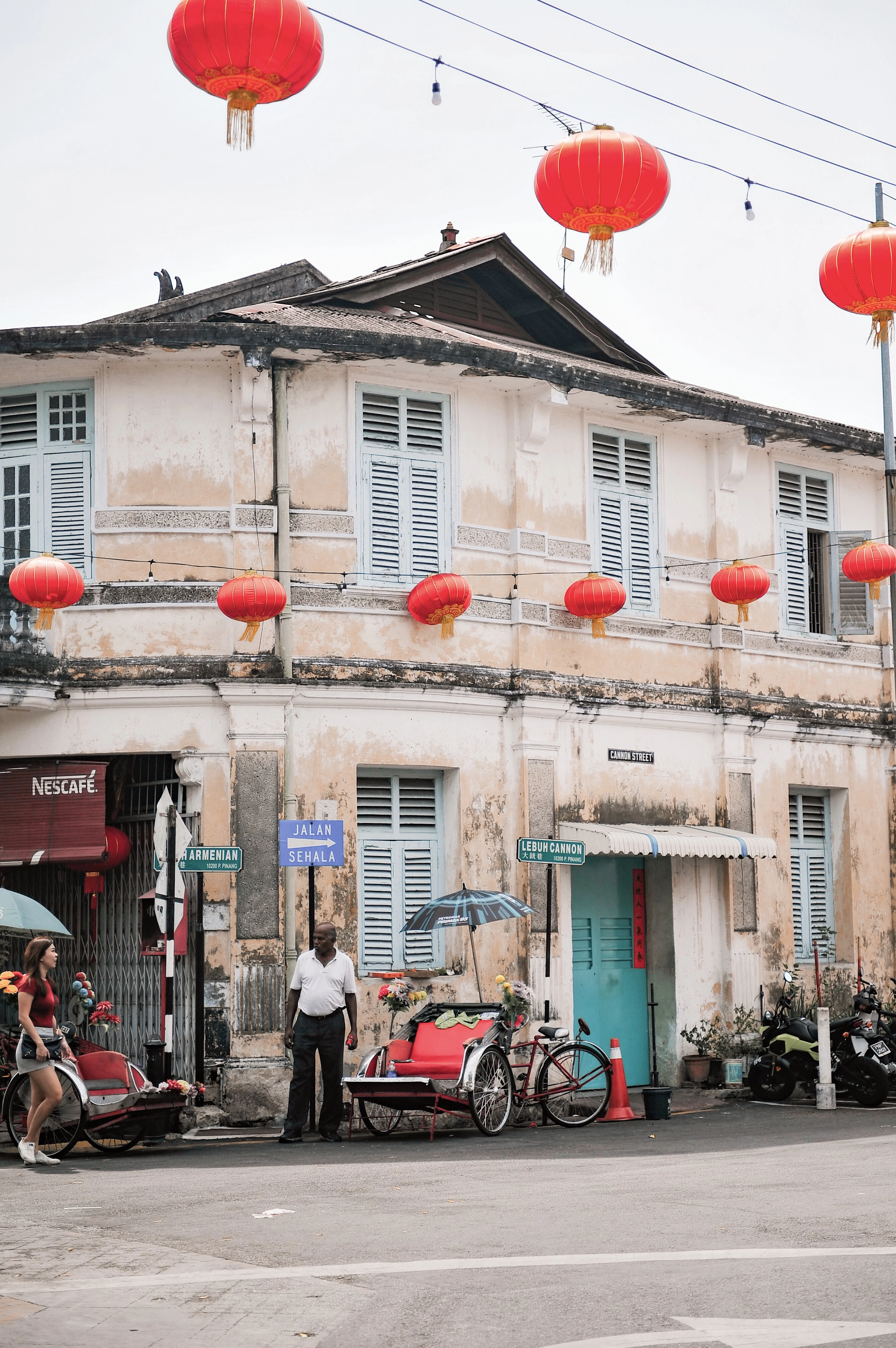 A weathered two-storey corner shophouse in the George Town heritage core, pale blue louvred shutters above peeling stucco and rows of red paper lanterns strung across the street at the junction of Armenian Street and Lebuh Cannon.