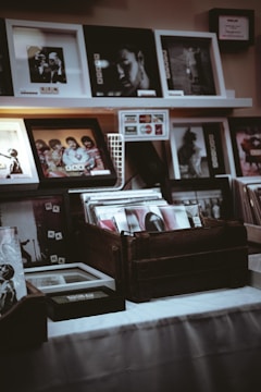 A collection of framed photographs and vinyl records displayed on shelves and wooden crates. Several photographs are in black and white, depicting portraits and candid shots. The lighting is dim, creating a vintage and nostalgic atmosphere.