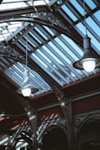 Two large pendant lights hang from an ornate and industrial-style ceiling featuring glass panels that allow natural light to illuminate the space. The structure includes intricate metalwork with a decorative pattern and a strong contrast between light and shadow.