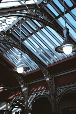 Two large pendant lights hang from an ornate and industrial-style ceiling featuring glass panels that allow natural light to illuminate the space. The structure includes intricate metalwork with a decorative pattern and a strong contrast between light and shadow.