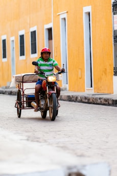 A person wearing a red helmet and green striped shirt rides a motorbike with a sidecar on a cobblestone street. The backdrop features a vibrant orange wall with several tall windows.