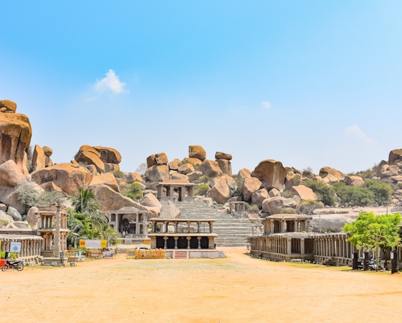 A historical site featuring ancient stone structures surrounded by large boulders. The architecture includes stone columns and steps leading up to more stone formations. Sparse vegetation, including a few trees and shrubs, is present.