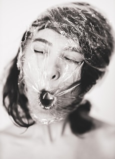 A close-up of a person's face wrapped in clear plastic wrap. The person's eyes are closed, and their mouth is open as if they are gasping for air. The image is in black and white, highlighting the texture of the plastic wrap and the contrast between the smooth skin and the wrapping material.