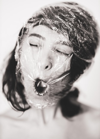 A close-up of a person's face wrapped in clear plastic wrap. The person's eyes are closed, and their mouth is open as if they are gasping for air. The image is in black and white, highlighting the texture of the plastic wrap and the contrast between the smooth skin and the wrapping material.