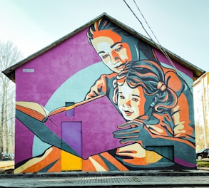 A large mural adorns the side of a house, depicting an adult embracing a child as they read a book together. The artwork uses a vibrant color palette and stylized, bold lines to create an engaging scene. The figures are depicted in warm and cool tones of orange, pink, and blue against a purple backdrop, creating a lively and inviting atmosphere.