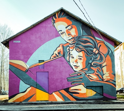 A large mural adorns the side of a house, depicting an adult embracing a child as they read a book together. The artwork uses a vibrant color palette and stylized, bold lines to create an engaging scene. The figures are depicted in warm and cool tones of orange, pink, and blue against a purple backdrop, creating a lively and inviting atmosphere.