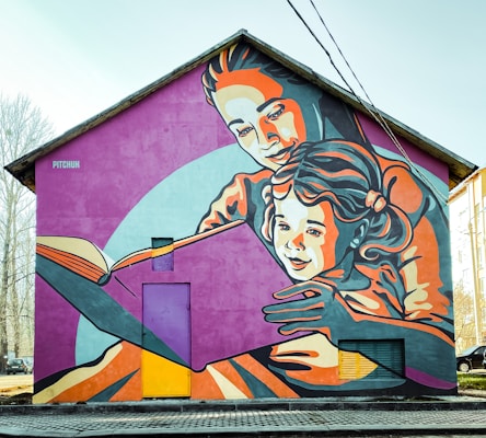 A large mural adorns the side of a house, depicting an adult embracing a child as they read a book together. The artwork uses a vibrant color palette and stylized, bold lines to create an engaging scene. The figures are depicted in warm and cool tones of orange, pink, and blue against a purple backdrop, creating a lively and inviting atmosphere.