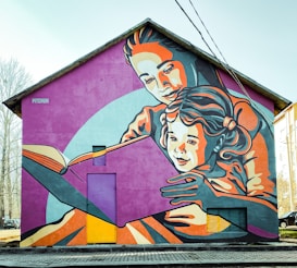 A large mural adorns the side of a house, depicting an adult embracing a child as they read a book together. The artwork uses a vibrant color palette and stylized, bold lines to create an engaging scene. The figures are depicted in warm and cool tones of orange, pink, and blue against a purple backdrop, creating a lively and inviting atmosphere.
