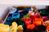A basket filled with neatly arranged rolls of colorful felt fabric. The fabrics are rolled tightly and organized by color, creating a vibrant and visually appealing display. Shades range from blues and greens to reds, oranges, and yellows.