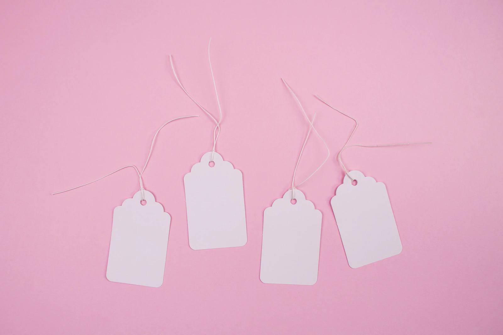 Four blank white paper price tags on strings, laid on a pale pink background.